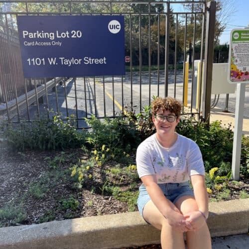 Student sitting in front of UIC Parking Lot 20, located at 1101 W. Taylor Street, with a sign indicating card access only. Nearby is a sign for the Monarch Memorial Garden, highlighting UIC's commitment to sustainability.