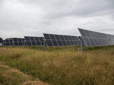 A solar panel farm featuring multiple rows of solar panels installed on metal frames, set against a backdrop of cloudy skies and tall grass.