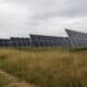 A solar panel farm featuring multiple rows of solar panels installed on metal frames, set against a backdrop of cloudy skies and tall grass.
